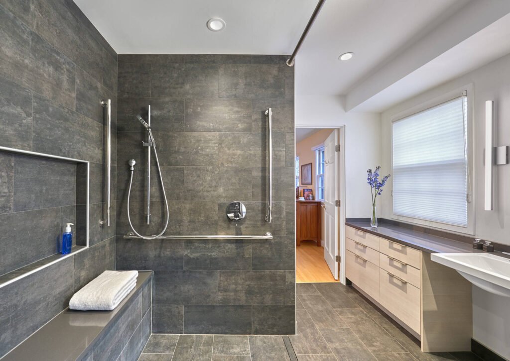 Modern walk-in shower with teak bench, matte black grab bars, and textured grey tiles